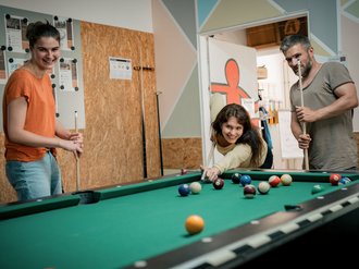 Drei junge Menschen spielen gemeinsam Billard. Eine Frau beugt sich über den grünen Tisch, um einen Stoß auszuführen, während eine andere Frau und ein Mann mit Queues daneben stehen und das Spiel beobachten.