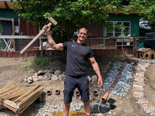 Ein lächelnder Mann in dunkler Kleidung steht im Freien und hält einen großen Holzhammer in die Höhe. Im Hintergrund ist eine Baustelle mit Rohren, Steinen und einem kleinen Schuppen zu sehen.