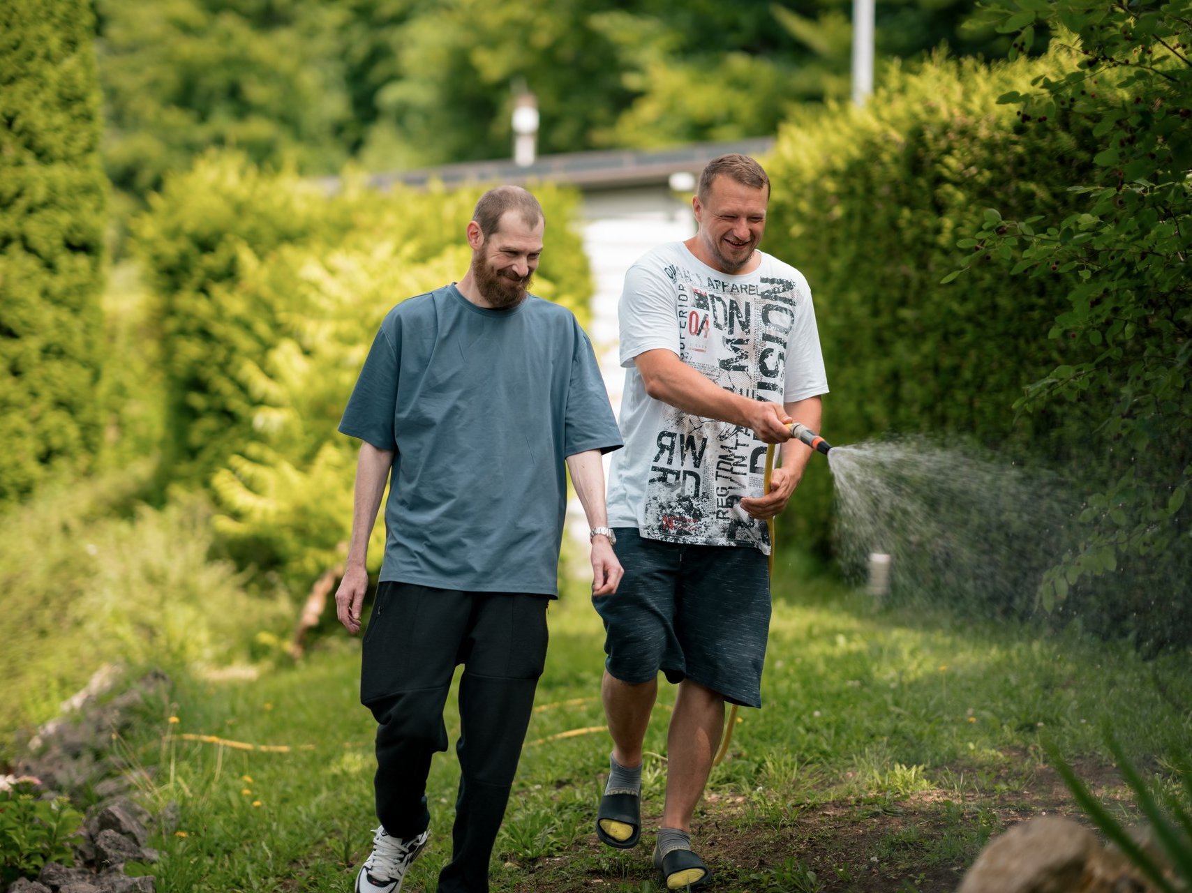 Zwei Männer gießen mit einem Gartenschlauch Blumen.