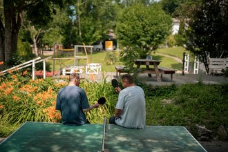 Zwei Männer sitzen von hinten auf einem grünen Tischtennistisch im Freien. Sie halten Tischtennisschläger in den Händen. Im Hintergrund ist eine grüne Parklandschaft mit Bäumen, Blumen und einem Picknicktisch zu sehen.