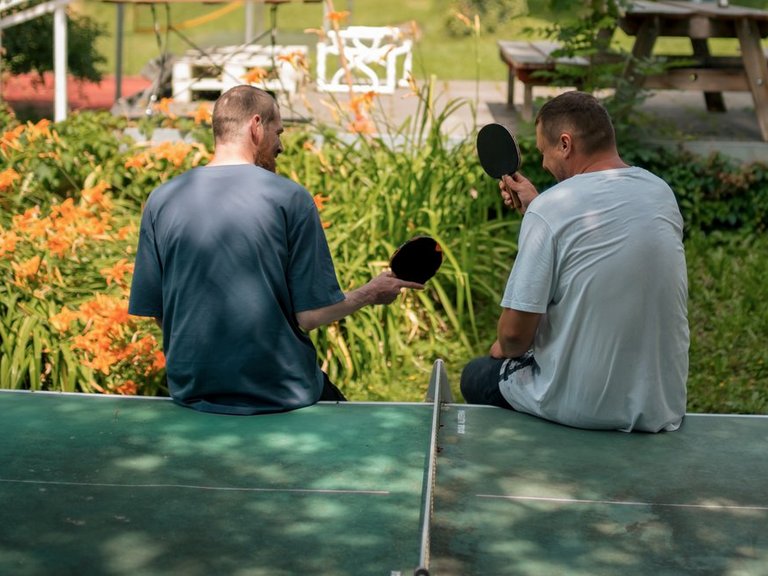 Zwei Männer sitzen von hinten auf einem grünen Tischtennistisch im Freien. Sie halten Tischtennisschläger in den Händen. Im Hintergrund ist eine grüne Parklandschaft mit Bäumen, Blumen und einem Picknicktisch zu sehen.