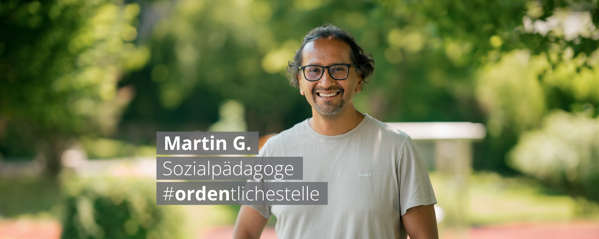 Suchthilfe_Fachklinik Grafrath_München_Team_Karriere Porträt von Martin G., einem lächelnden Mann mit Brille und grauem T-Shirt im Freien. Ein Textoverlay nennt seinen Beruf Sozialpädagoge und den Hashtag #ordentlichestelle.