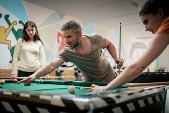 Drei Personen spielen in einem Spielzimmer Billard. Ein Mann mit grauem Bart zielt konzentriert mit dem Queue, während eine Frau rechts ebenfalls am Tisch steht und eine andere Frau im Hintergrund zuschaut.