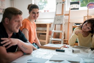 Drei Personen sitzen in einem hellen Atelier um einen Tisch mit Kunstmaterialien. Eine Frau in einem orangefarbenen Shirt und eine in Gelb lächeln fröhlich, während ein Mann links zuhört. Auf dem Tisch liegen Druckvorlagen und eine Walze.