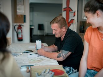 Ein Mann mit einem Armtattoo in einem schwarzen T-Shirt sitzt an einem Tisch und hält ein Smartphone. Eine Frau in einem orangefarbenen Shirt sitzt neben ihm, schaut auf das Handy und lächelt. Auf dem Tisch liegen Stifte und Papier.