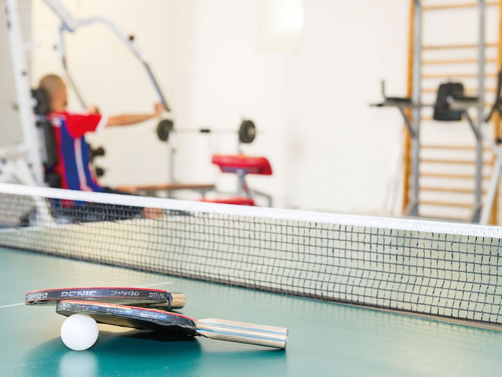 auf einer Tischtennisplatte liegen zwei Schläger und ein Ball - im Hintergrund trainiert jemand