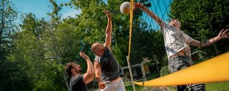 Drei Männer spielen Volleyball auf einem roten Außenplatz. Ein Spieler springt hoch, um den Ball über das Netz zu schlagen, während ein Gegenspieler versucht zu blocken. Im Hintergrund sind grüne Bäume und ein blauer Himmel zu sehen.