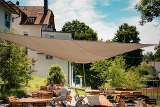 Zwei Männer unterhalten sich an einem Holztisch auf einer sonnigen Terrasse. Ein großes Sonnensegel spendet Schatten. Im Hintergrund sind ein Haus und üppiges Grün zu sehen.