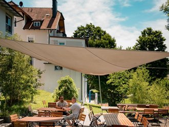 Zwei Männer unterhalten sich an einem Holztisch auf einer sonnigen Terrasse. Ein großes Sonnensegel spendet Schatten. Im Hintergrund sind ein Haus und üppiges Grün zu sehen.