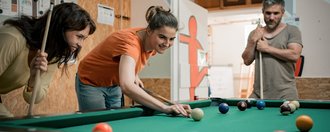 Zwei junge Frauen spielen Billard. Eine Frau im orangenen Shirt beugt sich zum Abstoß über den Tisch, während eine Freundin daneben steht und lächelnd zuschaut. Im Hintergrund beobachtet ein Mann das Spiel. Auf dem grünen Tuch liegen mehrere farbige Billardbälle.