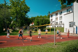 Eine Gruppe von Menschen spielt Volleyball auf einem roten Außenplatz. Ein gelbes Netz teilt das Spielfeld. Im Hintergrund sind grüne Bäume und ein weißes Gebäude unter blauem Himmel zu sehen.
