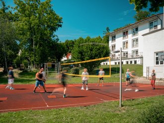 Eine Gruppe von Menschen spielt Volleyball auf einem roten Außenplatz. Ein gelbes Netz teilt das Spielfeld. Im Hintergrund sind grüne Bäume und ein weißes Gebäude unter blauem Himmel zu sehen.