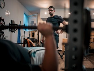 Ein Mann führt eine Bankdrück-Übung mit einer Langhantel aus, während ein Trainer mit Brille im Hintergrund aufmerksam zuschaut.