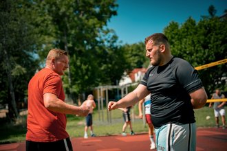 Zwei Männer stehen sich auf einem Sportplatz gegenüber. Der Mann im orangefarbenen T-Shirt und der Mann im schwarzen T-Shirt heben ihre Fäuste, als wollten sie sich die Fäuste stoßen. Im Hintergrund sind unscharf weitere Personen und Bäume zu erkennen.