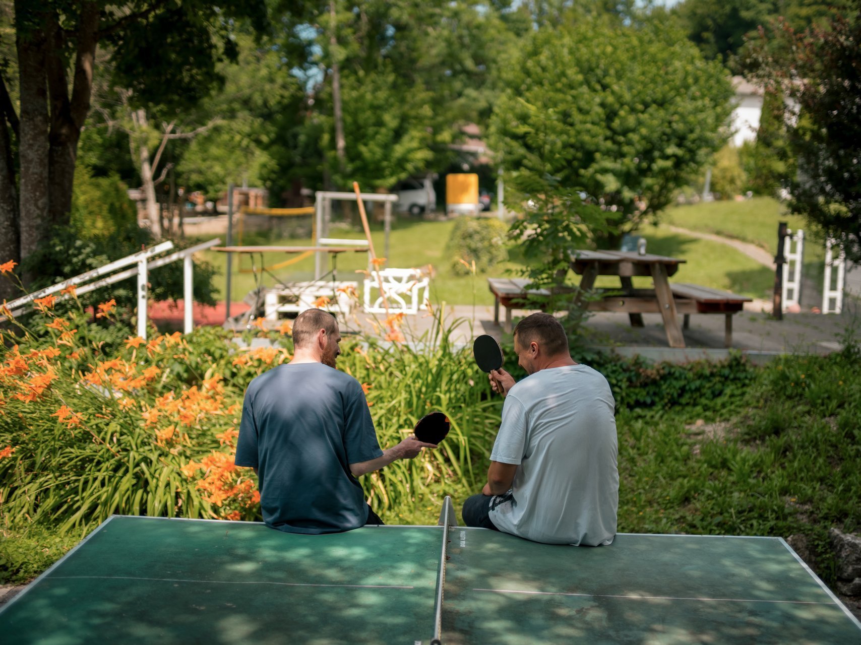 Zwei Männer lehnen mit dem Rücken zur Kamera an einer Tischtennisplatte in einem Garten und unterhalten sich.
