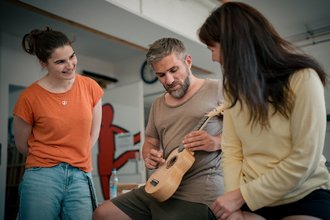 Drei Personen sitzen im Kreis. Ein Mann in der Mitte spielt Ukulele. Eine Frau links im orangen T-Shirt und eine Frau rechts im gelben Oberteil schauen ihm lächelnd zu.