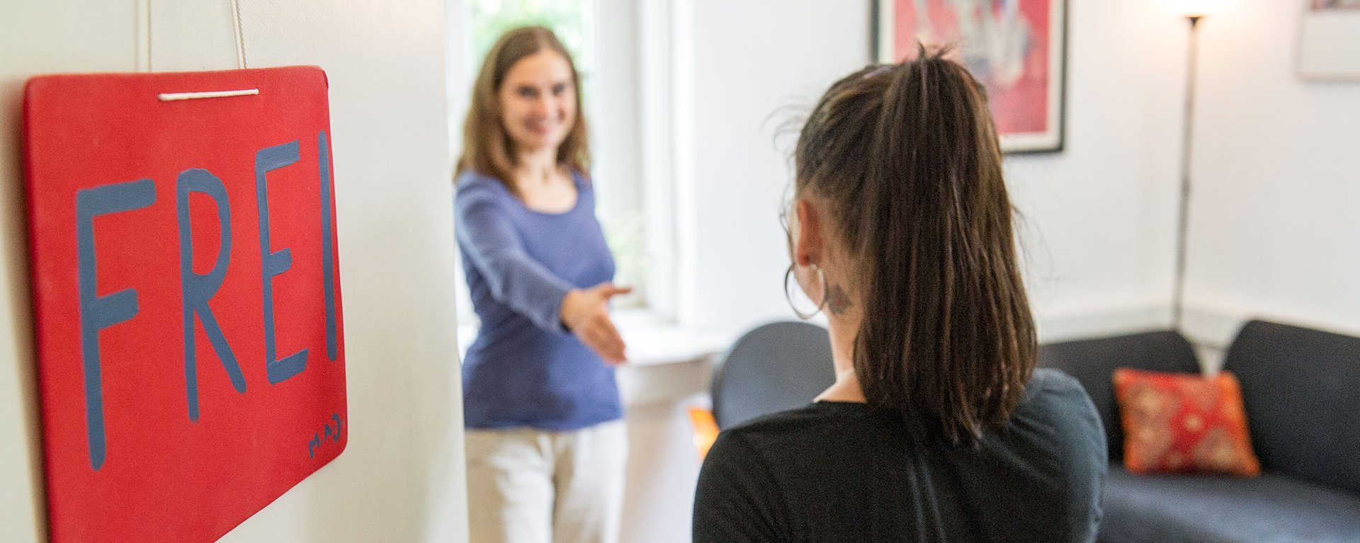 Eingangstüre mit einem, in rot, gemalten Schild und der Aufschrift: "FREI". Im Hintergrund begrüßen sich zwei Frauen