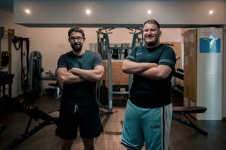 Zwei lächelnde Männer stehen mit verschränkten Armen in einem Fitnessstudio. Der Mann links trägt eine Brille und schwarze Sportkleidung, während der Mann rechts ein dunkelgrünes Shirt und hellblaue Shorts trägt. Im Hintergrund sind Trainingsgeräte zu erkennen.