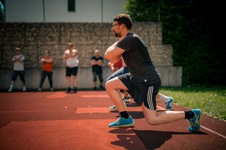 Ein Mann in sportlicher Kleidung führt auf einer roten Laufbahn einen Ausfallschritt aus. Er trägt blaue Turnschuhe und eine Brille. Im unscharfen Hintergrund stehen mehrere andere Personen vor einer Steinmauer und beobachten das Training.