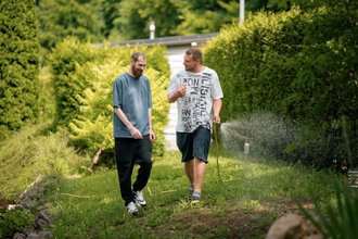 Zwei Männer gehen durch einen grünen Garten. Der Mann rechts hält einen Gartenschlauch und gießt die Pflanzen, während der Mann links neben ihm herläuft und lächelt.