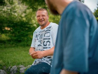 Ein Mann mit Bart und weißem T-Shirt sitzt im Grünen und lacht herzlich. Er blickt auf eine Person im Vordergrund, deren Schulter unscharf zu sehen ist. Die Stimmung ist fröhlich.
