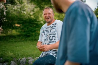 Ein Mann mit Bart und weißem T-Shirt sitzt im Grünen und lacht herzlich. Er blickt auf eine Person im Vordergrund, deren Schulter unscharf zu sehen ist. Die Stimmung ist fröhlich.
