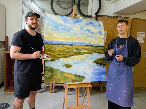 Zwei Männer stehen in einem Atelier auf beiden Seiten eines großen Landschaftsgemäldes auf einem Staffelei. Das Ölgemälde zeigt einen Fluss, grüne Wiesen und Berge im Hintergrund. Der Mann links hält eine Farbpalette, der Mann rechts trägt eine blaue Schürze.