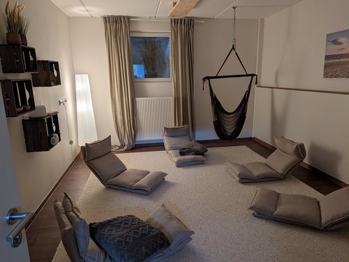 Ein entspannungsfreundlicher Raum mit mehreren grauen Bodensesseln, die kreisförmig auf einem Teppich angeordnet sind. Ein schwarzer Hängesessel hängt an der Decke. An der Wand sind Regale mit Pflanzen und Lautsprechern zu sehen.
