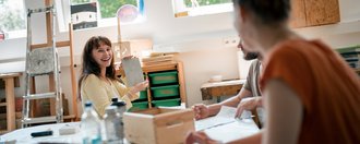 Eine glückliche Frau in einem gelben Oberteil sitzt an einem Tisch in einem hellen Atelier und hält eine Skizze hoch, um sie zwei anderen Personen zu zeigen. Im Hintergrund sind Staffeleien und Kunstmaterialien zu erkennen.