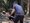 Ein Mann in grauem T-Shirt und schwarzen Shorts schwingt einen großen Schlaghammer im Wald. Im Hintergrund liegt ein schwarz-weißer Hund entspannt auf dem Waldboden.