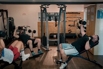 Drei Männer liegen auf Schrägbänken in einem Fitnessstudio und führen eine Brustpresse mit Kurzhanteln aus. Im Hintergrund sind weitere Trainingsgeräte wie ein Kabelzuggerät zu sehen.