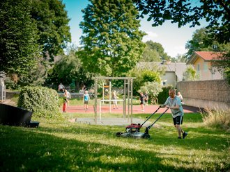 Ein Mann mäht im Vordergrund einen grünen Rasen. Im unscharfen Hintergrund ist ein Sportplatz mit rotem Boden zu sehen, auf dem Kinder und Jugendliche ein Ballspiel ausüben. Die Szene ist sonnig.