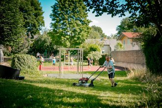 Ein Mann mäht im Vordergrund einen grünen Rasen. Im unscharfen Hintergrund ist ein Sportplatz mit rotem Boden zu sehen, auf dem Kinder und Jugendliche ein Ballspiel ausüben. Die Szene ist sonnig.