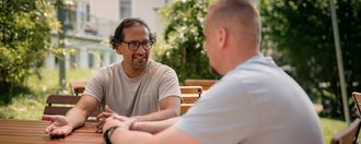 Zwei Männer unterhalten sich an einem Holztisch im Freien. Der Mann mit Brille und beigem T-Shirt gestikuliert beim Sprechen, während ihm sein Gesprächspartner zuhört. Im Hintergrund sind grüne Bäume und ein Haus zu sehen.