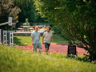Zwei Männer gehen nebeneinander auf einem roten Weg in einem grünen Garten. Im Hintergrund sind Bäume und ein Basketballkorb sichtbar. Sie tragen legere Kleidung und scheinen sich zu unterhalten.