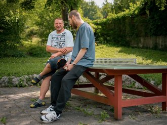 Zwei Männer sitzen lächelnd auf einem roten Tischtennistisch im Freien und unterhalten sich. Sie befinden sich in einer grünen Parkanlage mit Bäumen im Hintergrund.