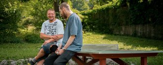 Zwei Männer lehnen an einer Tischtennisplatte und unterhalten sich.