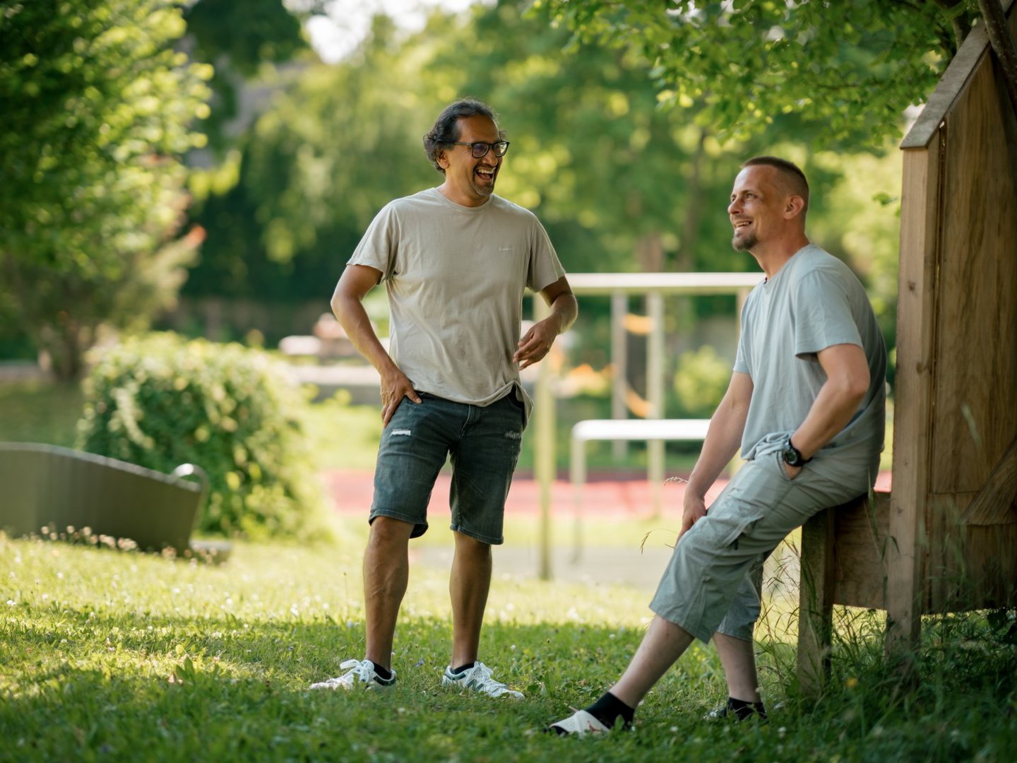 Zwei Männer unterhalten sich fröhlich auf einer Wiese im Park. Einer steht, der andere sitzt an einem Holzgebäude. Beide lächeln und wirken entspannt bei sonnigem Wetter.