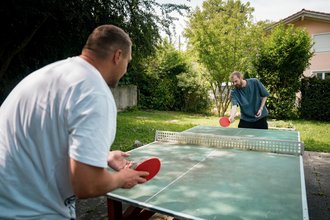 Zwei Männer spielen Tischtennis im Garten. Der Mann im Vordergrund trägt ein weißes T-Shirt, sein Gegenspieler gegenüber ein blaues. Beide halten rote Schläger und konzentrieren sich auf das Spiel am grünen Tischtennistisch.
