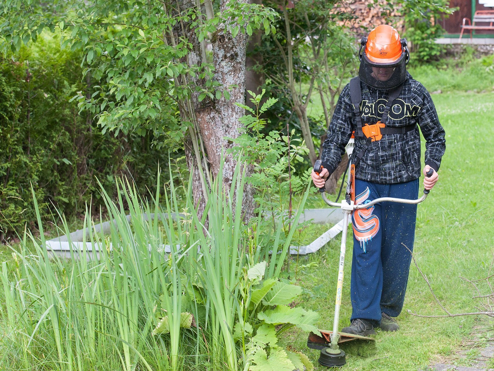 Gartenarbeit: ein Mann mit Arbeitssicherheits-Ausrüstung und orangem Helm trimmt den Rasen