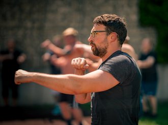 Ein Mann mit Bart und Brille trainiert Boxen im Freien. Er führt einen Schlag mit dem linken Arm aus, während im unscharfen Hintergrund andere Teilnehmer ebenfalls Übungen durchführen.