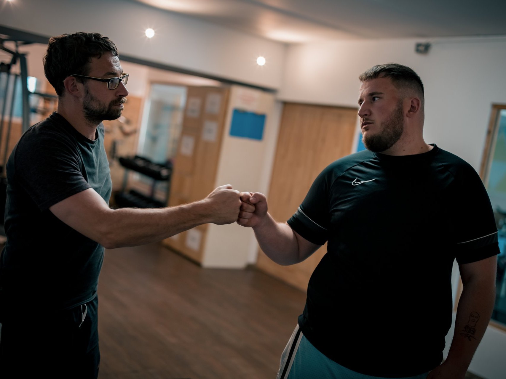 Zwei Männer in einem Fitnessstudio geben sich zur Begrüßung die Faust. Beide tragen dunkle T-Shirts. Der Mann links trägt eine Brille. Die Geste symbolisiert Motivation und Zusammenhalt.