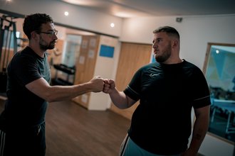 Zwei Männer in einem Fitnessstudio geben sich zur Begrüßung die Faust. Beide tragen dunkle T-Shirts. Der Mann links trägt eine Brille. Die Geste symbolisiert Motivation und Zusammenhalt.